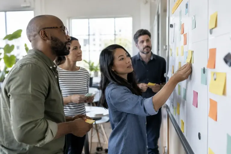 A small team moving unlettered task cards across a planning board during a practical meeting.