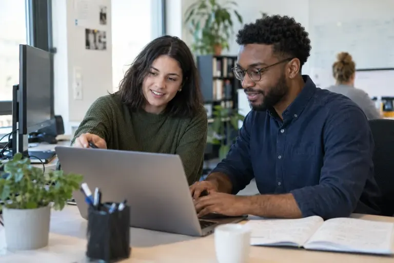 New employee and colleague working through a small practical task together at an ordinary workstation, friendly professional scene.